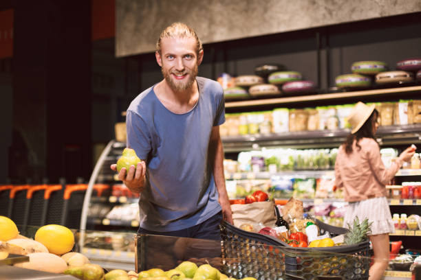 istock Young smiling man joyfully looking in camera holding pear in hand with trolley full of products near while pretty girl on background choosing products in modern supermarket 1357286536