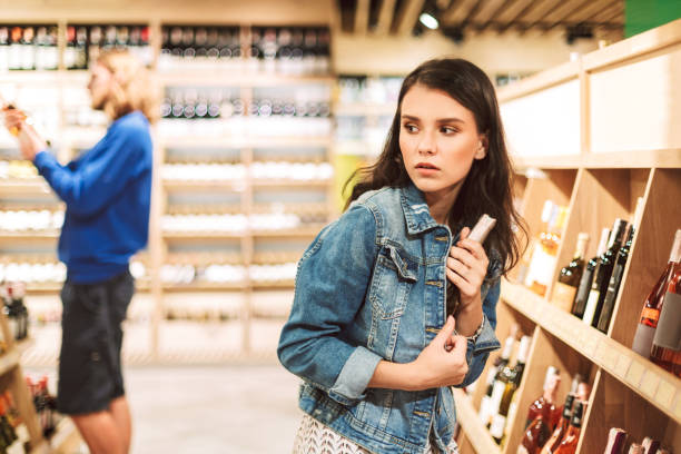 現代のスーパーマーケットでワインのボトルを盗もうとしているデニムジャケットの若い恐ろしい女性 - wine wine bottle drink alcohol ストックフォトと画像