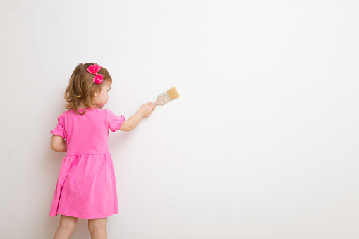 Baby girl in pink dress holding new paint brush and coloring white wall background at nursery room. Empty place for text.