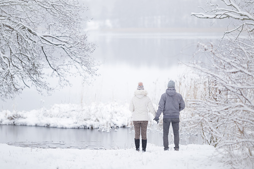 Young couple standing on snow at lake shore and holding each other hands and looking far away. Romantic lovely moment at beautiful place. Peaceful atmosphere. White cold snowy winter day. Back view.
