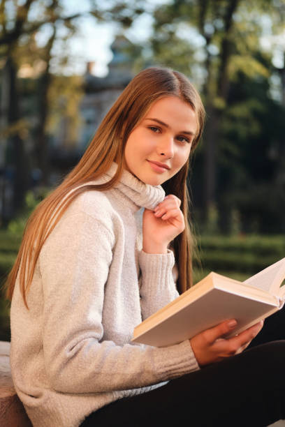 istock Young pretty smiling student girl in cozy sweater sensually looking in camera studying with book outdoor 1357301992