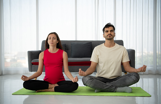 A pregnant woman meditates with her husband in a living room. by practicing meditation in serene harmony in lotus position