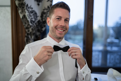 happy bridegroom smiling at camera while putting on his bow tie during wedding preparations