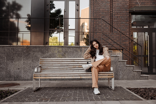 Tall stylish girl with oversized jacket sitting on the bench of the city background. High quality photo