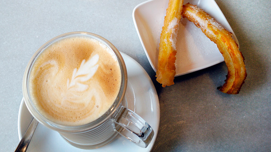 Close up of a white cup of cappuccino and two churros. Selective focus on the foreground,  high angle view.