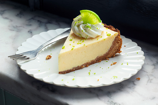 A slice of creamy key lime pie topped with a garnish of whipped cream and lime zest, with a small citrus slice. The crust is made from graham crackers or cookies and the piece of pie is on a white plate with a fork on a marble counter top.
