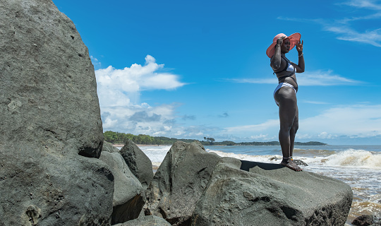 African woman with sun hat standing on the rocky cliffs at the tropical beach in Axim Ghana West Africa
