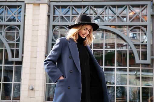 Lifestyle portrait of fashionable woman wearing winter or spring outfit, felt hat, gray wool coat and turtleneck. Outdoors. Female stylish Model smiling, walking city Street. Street fashion trend