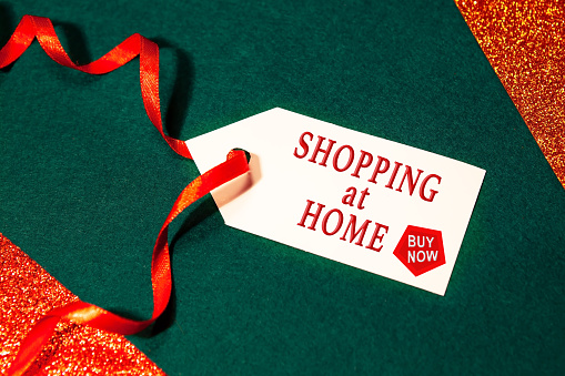 Label with the text - shopping at home, buy now, with a red ribbon, on a green background. The concept of safety during the coronavirus pandemic on New Year's holidays.