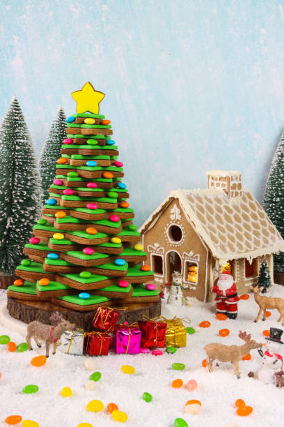 Image of homemade, gingerbread house and Christmas tree made from a stack of star shaped gingerbread biscuits decorated with royal icing displayed in snowy conifer forest scene, model fir trees, icing sugar snow, snowy blue background
