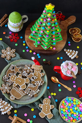 Stock photo showing close-up view of wooden chopping board containing a stack of star shaped gingerbread biscuits, decorated with green royal icing and multi-coloured candy coated chocolate sweets, forming a Christmas tree. Home baking concept.