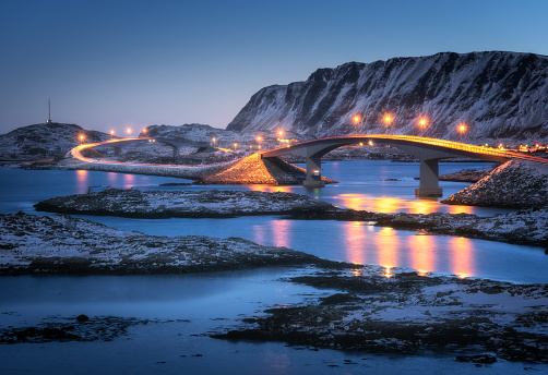 Bridge with illumination, snow covered mountains, village and blue sky with reflection in water. Night landscape with bridge, snowy rocks, city lights, sea. Winter in Lofoten islands, Norway. Road