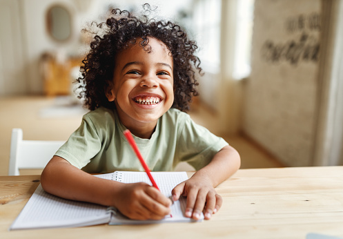 Niño afroamericano sonriente haciendo la tarea mientras está sentado en el escritorio de casa photo
