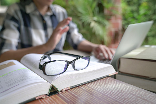 Books on table with woman on online video call at the background. Selective focus on eyeglasses.