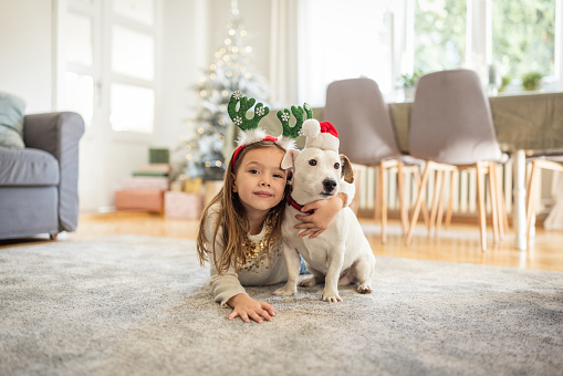 Young girl with pet dog sitting in front of Christmas tree at home.