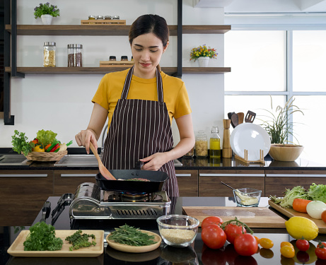 Young asian woman dressed in an apron cooking steak. The kitchen counter full of various kinds of vegetables and seasonings.
