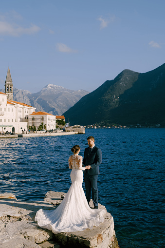 Groom and bride stand on a stone pier against the background of the ancient town of Perast. High quality photo