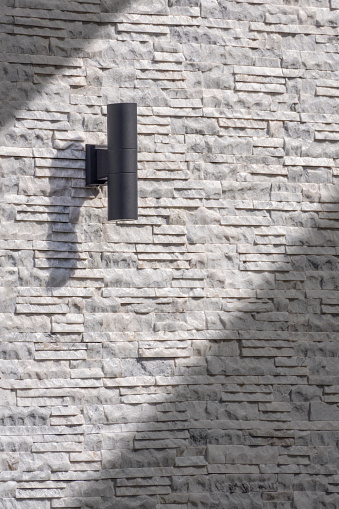 Sunlight and shadow on surface of outdoor black cylinder wall lamp on vintage white and gray ledgestone wall in vertical frame