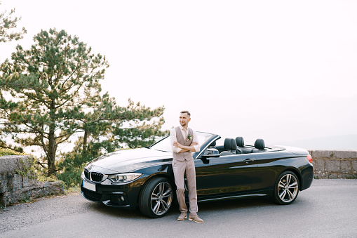 Groom is leaning against the convertible on the road. High quality photo