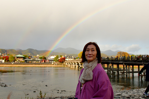 A Japanese woman in her early 60’s is enjoying autumn leaf color of Arashiyama, Kyoto, on a foggy, rainy day, with double rainbow behind her.   
Arashiyama is located in the Western outskirts of Kyoto and a popular destination for tourists as well as Kyoto people for its beautiful natural settings, especially during cherry blossom and autumn color seasons.