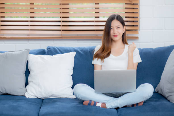 Young asian business woman smile and work from home with laptop computer online to internet on sofa in living room, freelance girl using notebook on couch with comfort, new normal, lifestyle concept. joven mujer de negocios asiática sonríe y trabaja desde casa con computadora portátil en línea a internet en el sofá en la sala de estar, chica independiente que usa el cuaderno en el sofá con comodidad, nueva normalidad, concepto de estilo de vida.