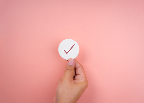 Checklist concept, minimal style. Check mark icon symbols on white round sponge in hand on pink pastel background.