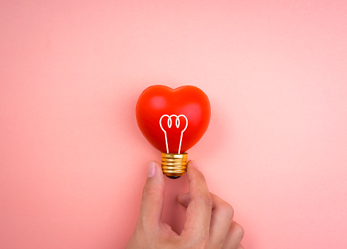 Hand holding light bulb icon symbol on red heart ball on pink pastel background, minimal style. Love, care, sharing, giving, wellbeing, inspiration, and idea concepts.