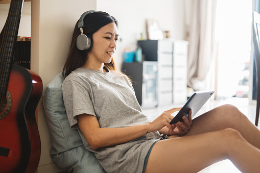 Shot of a happy Asian woman relaxing sitting on her living room floor and using headphones with a digital tablet.
