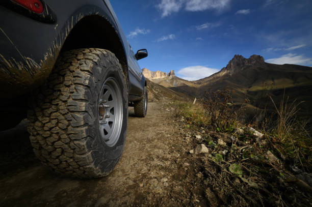 Close-up A large off-road all-terrain car wheel stands on a rough road in the mountains against the backdrop of rocks. Off-road tours and off-road travel concept stock photo