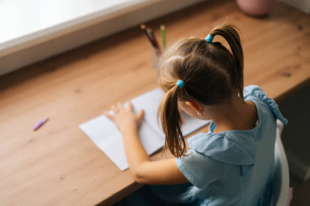 istock High-angle view of unrecognizable little child girl learning writing doing homework sitting at home table by window. 1357406440