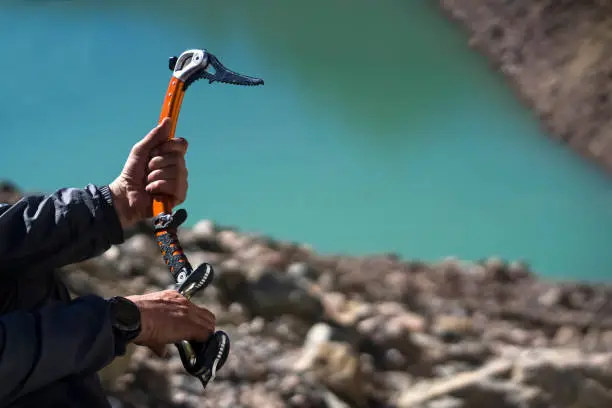 Male hands hold an ice ax on the background of a mountain lake from a glacier. A man takes ice tools while climbing the mountain.