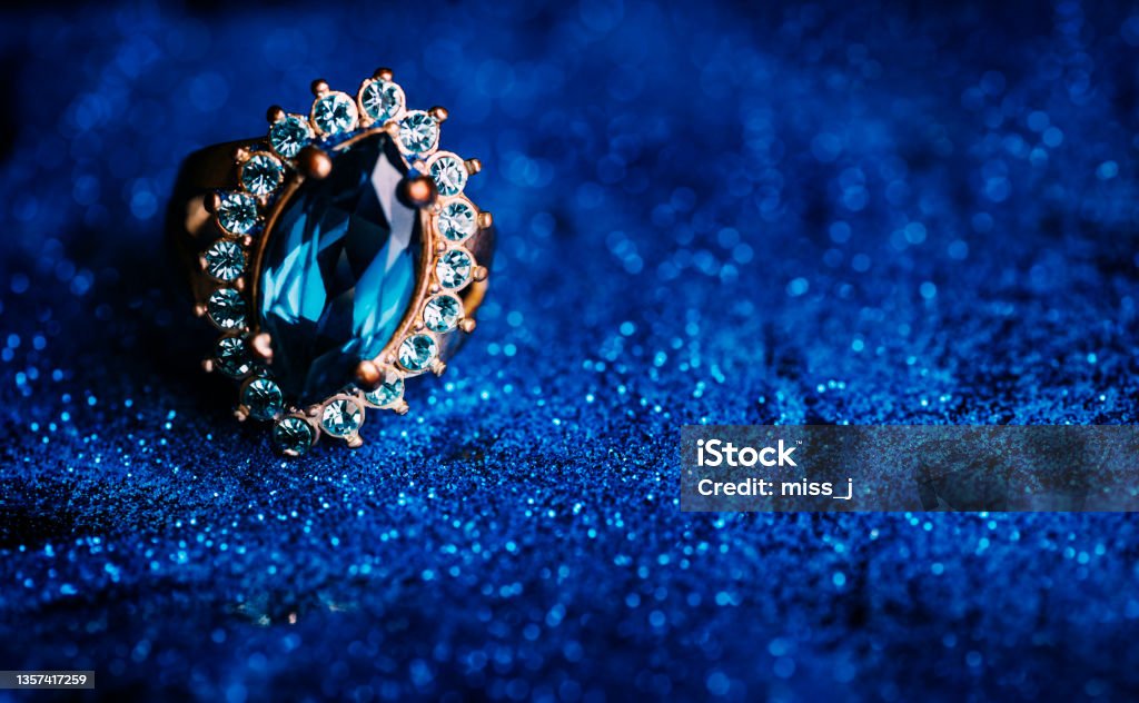 Closeup of luxury wedding ring in dark blue glitter background. Closeup of luxury wedding ring in dark blue glitter background. Bridal golden jewelry gift with precious gemstone. Jewelry Stock Photo