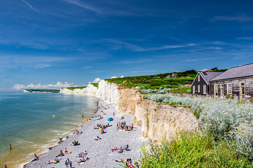 Color image depicting a senior man in his 70s hiking on a path leading down to the iconic Seven Sisters cliffs on the coastline of East Sussex, UK. The man is wearing casual clothing - blue denim jeans, a red checked shirt, navy blue gilet and a rucksack.