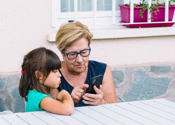 Grandmother and granddaughter with surprised faces looking at the mobile on a terrace. Family, grandchildren, grandparents and technology concept. stock photo
