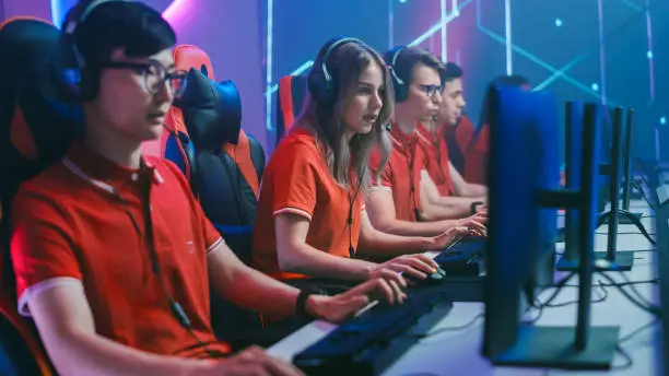 Photo of Diverse Esport Team of Pro Gamers Play in Mock-up RPG Strategy Video Game on a Championship. Stylish Neon Cyber Games Arena. Online Broadcasting of Tournament Event. Side View Shot