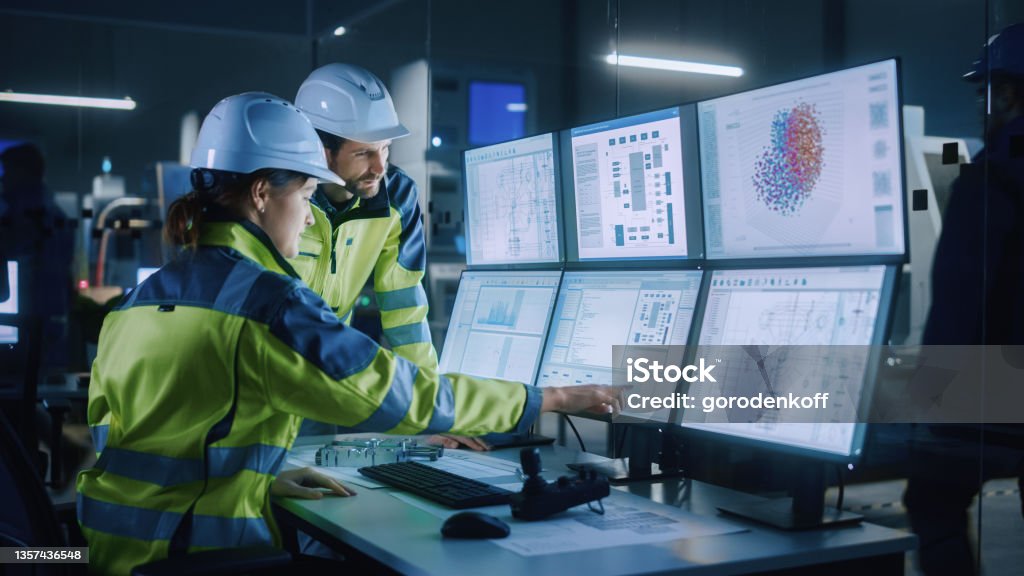 Industry 4.0 Modern Factory: Project Engineer Talks to Female Operator who Controls Facility Production Line, Uses Computer with Screens Showing AI, Machine Learning Enhanced Assembly Process Factory Stock Photo
