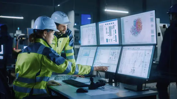 Photo of Industry 4.0 Modern Factory: Project Engineer Talks to Female Operator who Controls Facility Production Line, Uses Computer with Screens Showing AI, Machine Learning Enhanced Assembly Process