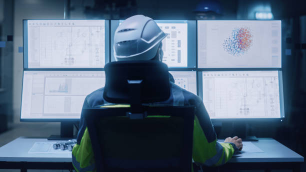 istock Industry 4.0 Modern Factory: Facility Operator Wearing Hardhat and Safety Vest Controls Workshop Production Line, Uses Computer with Screens Showing Machinery Control Operation UI. Back View Shot 1357436615