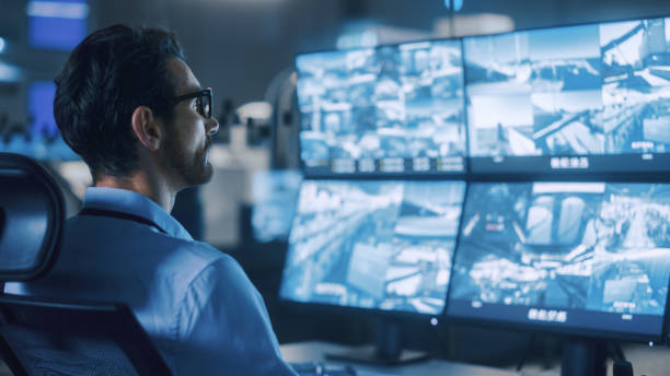 Industry 4.0 Modern Factory: Portrait Shot of a Security Operator Controls Proper Functioning of Workshop Production Line, Uses Computer with Screens Showing Surveillance Feed. High-Tech Security industria 4.0 fábrica moderna: toma de retrato de un operador de seguridad controla el correcto funcionamiento de la línea de producción del taller, utiliza una computadora con pantallas que muestran la alimentación de vigilancia. seguridad de alta tec