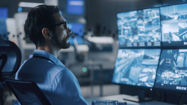 istock Industry 4.0 Modern Factory: Portrait Shot of a Security Operator Controls Proper Functioning of Workshop Production Line, Uses Computer with Screens Showing Surveillance Feed. High-Tech Security 1357436634