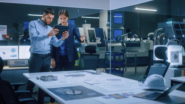 istock Factory Office Meeting Room: Male Chief Engineer Holds Digital Tablet Computer, Shows Screen to Female Project Manager, They Talk, Find Optimization Solution. Modern Industry 4.0 CNC Machinery 1357436645