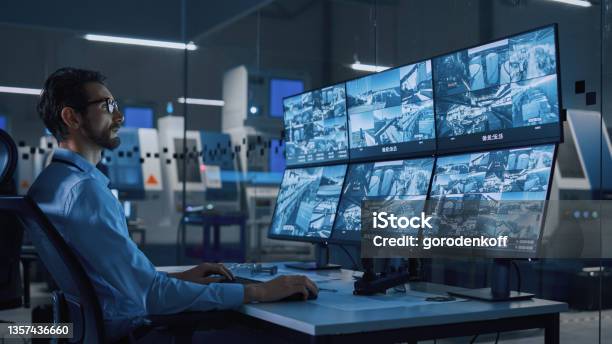 Industry 40 Modern Factory Security Operator Controls Proper Functioning Of Workshop Production Line Uses Computer With Screens Showing Surveillance Camera Feed Hightech Security Stock Photo - Download Image Now