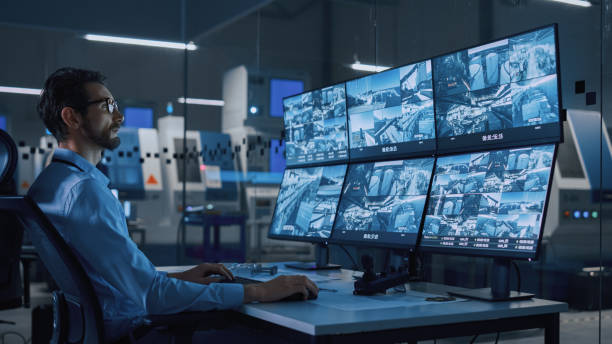 industry 4.0 modern factory: security operator controls proper functioning of workshop production line, uses computer with screens showing surveillance camera feed. high-tech security - espião imagens e fotografias de stock