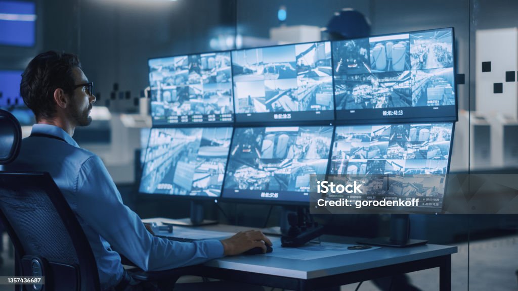Industry 4.0 Modern Factory: Security Operator Controls Proper Functioning of Workshop Production Line, Uses Computer with Screens Showing Surveillance Camera Feed. High-Tech Security Security Stock Photo