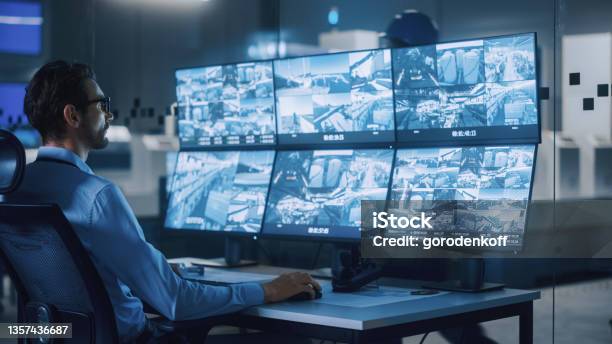 Industry 40 Modern Factory Security Operator Controls Proper Functioning Of Workshop Production Line Uses Computer With Screens Showing Surveillance Camera Feed Hightech Security Stock Photo - Download Image Now