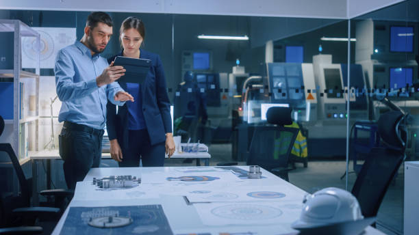 istock Factory Office Meeting Room: Male Chief Engineer Holds Digital Tablet Computer, Shows Screen to Female Project Manager, They Talk, Find Optimization Solution. Modern Industry 4.0 CNC Machinery 1357436689