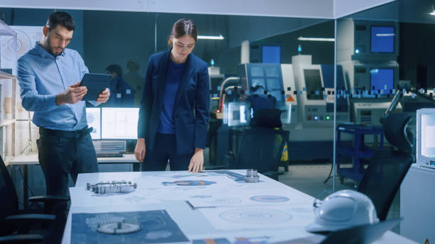 istock Factory Office Meeting Room: Male Chief Engineer Holds Digital Tablet Computer, Shows Screen to Female Project Manager, They Talk, Find Optimization Solution. Modern Industry 4.0 CNC Machinery 1357436716