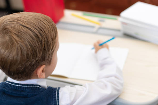 istock A cute boy first grader in a school uniform does homework while sitting at a desk with a pencil in his hands at home. Remote education. Selective focus. Close-up 1357444351