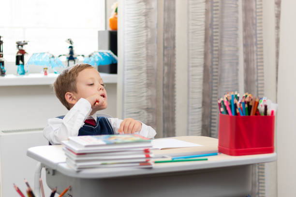 istock A cute boy first grader in a school uniform does homework while sitting at a desk with a pencil in his hands at home. Remote education. Selective focus. Close-up 1357444352