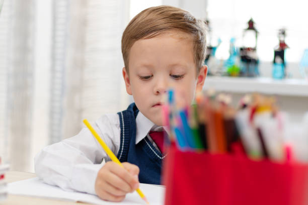 istock A cute boy first grader in a school uniform does homework while sitting at a desk with a pencil in his hands at home. Remote education. Selective focus. Close-up 1357444365
